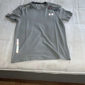 Grå Under Armour t-shirt - Snygg grå t-shirt från Under Armour med logga på bröstet och färgglad text längs sidan. T-shirten har korta ärmar och är tillverkad i ett lätt och sportigt material som passar perfekt till träning. Modern och stilren design med rund hals.