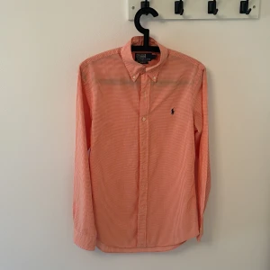 Polo Ralph Lauren skjorta  - Snygg orange rutig skjorta från Polo Ralph Lauren i slim fit. Klassisk button-down krage, långärmad och med små vita knappar framtill. Ikoniska Polo-loggan broderad på bröstet. Skjortan är gjord i mjuk bomull och passar perfekt till jeans eller chinos.