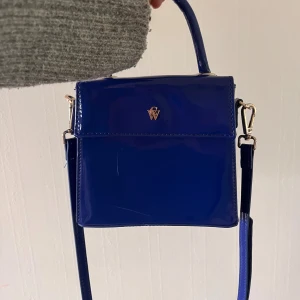 Fin blå handväska 💙 - Snygg blå axelväska i glansigt syntetmaterial från Carin Wester. Väskan har en axelrem som går att ta bort. Perfekt storlek för mobil och småsaker, med magnetlås och enkel logga framtill. Tyvärr några små skador på framsidan men inget som märks på håll💕💕