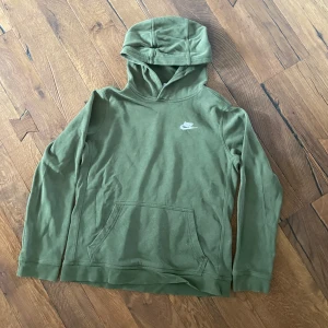 Olivgrön Nike hoodie  - Snygg olivgrön hoodie från Nike i storlek XL. Klassisk design med vit Nike-logga på bröstet, känguruficka och huva. Tillverkad i mjuk bomull, perfekt för chill dagar eller när du vill ha en avslappnad stil.