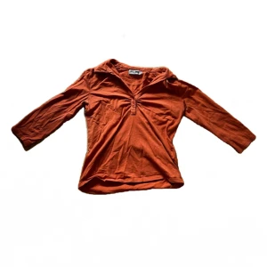 Orange topp från A feeling of Joy - Snygg orange topp från A feeling of Joy i mjuk bomull. Modellen har trekvartsärm, v-ringning med knappar och en avslappnad passform. Perfekt att styla med jeans eller kjol för en cool vardagslook.