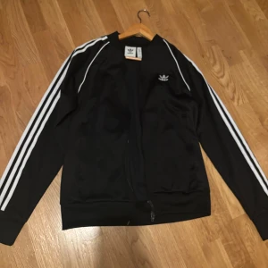 Svart Adidas track jacket med vita ränder - Svart Adidas track jacket med klassiska vita ränder längs ärmarna och broderad logga på bröstet. Jackan har dragkedja framtill och är tillverkad av polyester, vilket ger en sportig och clean look. Perfekt för dig som gillar streetwear och sportig stil.