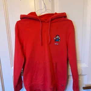 Röd Super Mario hoodie från Levi's XS - Röd hoodie från Levi's i samarbete med Super Mario. Framsidan har en broderad Mario och Levi's-logga, baksidan har texten 'It's-a me, Mario!'. Tröjan har huva med snörning och stor magficka.