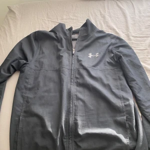 Svart vindjacka från Under Armour - Snygg svart vindjacka från Under Armour med dragkedja framtill och diskret logga på bröstet. Jackan har hög krage och är tillverkad i lätt polyester, perfekt för aktiva dagar eller när du vill ha en sportig look.