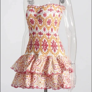 Vit, rosa, gul och röd kortklänning med volanger - Supersöt kortklänning i vitt med röda och gula mönster, strapless och med smockad överdel. Kjolen har två lager volanger med grafiskt tryck i rött och gult. Perfekt för dig som gillar färg och unika detaljer. Passar dig som vill sticka ut och ha något riktigt somrigt.