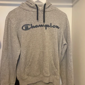 Grå hoodie från Champion med logga - Säljer en grå hoodie från Champion i storlek S. Tröjan har svart snörning i huvan, stor Champion-logga framtill och en klassisk känguruficka. Perfekt för chill dagar eller när du vill ha en sportig look.