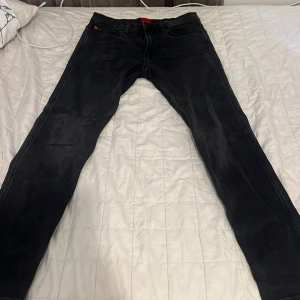 Svarta jeans från Hugo med röd patch - Svarta jeans från Hugo med en snygg röd patch med logga bak i midjan. Jeansen har klassisk femficksdesign, raka ben och diskreta slitningar för en cool look. Tillverkade i jeansmaterial med en mörk tvätt. Perfekta för dig som gillar stilrena och moderna byxor.