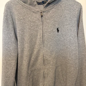 Grå hoodie från Polo Ralph Lauren - Gråmelerad hoodie från Polo Ralph Lauren med dragkedja framtill och klassisk huva. Tröjan har broderad svart logga på bröstet och känguruficka. Tillverkad i mjuk bomullsmix, perfekt för chill dagar eller när du vill ha en clean look.