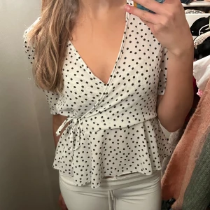 Vit omlottblus med svarta prickar - Supersöt vit omlottblus med svarta prickar och knyt i sidan. Blusen har korta ärmar, v-ringning och volang nedtill som ger en riktigt fin siluett. Perfekt för dig som gillar en lekfull och trendig stil.
