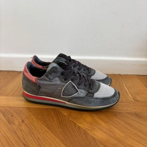 Grå sneakers från Philippe Model - Snygga sneakers från Philippe Model i grått med detaljer i rött och svart. Ovandelen är en mix av mocka, textil och skinn, med svarta snören och en vit/röd rand på sulan. Klassisk låg modell med rund tå och coola kontraster.