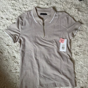 Beige piké med dragkedja från S.C.W. - Snygg beige piké från S.C.W. med vit kant vid krage och ärmslut. T-shirten har korta ärmar och en stilren half zip framtill. Helt ny aldrig använd