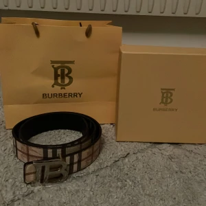Burberry rutiga bälten med B-spänne - Ett snyggt Burberry-bälten med klassiskt rutigt mönster och stort B-spänne i silver. Ett bälte i beige och bruna toner, det andra i svart och grått. Båda är i skinn och levereras med originalpåse och ask. Perfekt accessoar för att lyfta din outfit.