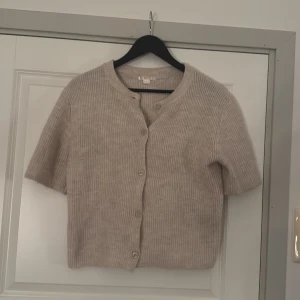 Kortärmad kofta  - Säljer en beige, ribbad kofta med korta ärmar och rund hals. Koftan har knappar framtill och är tillverkad i ett mjukt stickat material som känns skönt mot huden. Fick den i julas och har inte använt den så mycket, så den är i bra skick!