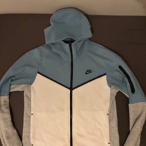 Nike hoodie i blått, vitt och grått M - Nike hoodie i snygg färgblock-design med ljusblå huva och överdel, vita paneler framtill och grå ärmar. Dragkedja hela vägen, svart Nike-logga på bröstet och praktisk ficka med dragkedja på ena ärmen. Perfekt för chill eller träning.