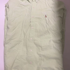 Ljusgrön skjorta från Ralph Lauren - Snygg ljusgrön skjorta från Ralph Lauren med klassisk krage och knappar hela vägen. Skjortan har den ikoniska broderade hästloggan på bröstet och är tillverkad i mjuk bomull. Perfekt för dig som gillar stilrena och tidlösa plagg.