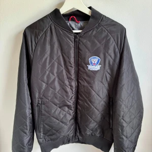 Svart quiltad bomberjacka Borås Hockey - Svart quiltad bomberjacka från ID Identity med Borås Hockey-logga på bröstet. Jackan har dragkedja framtill, ribbade muddar och två fickor med dragkedja. Snyggt rutmönster över hela jackan och klassisk bomberkrage. Perfekt för dig som vill sticka ut med sportig stil.