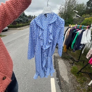 Blå prickig volangklänning Vero Moda - Säljer en blå klänning från Vero Moda med vita prickar, volangdetaljer längs halsringningen och nederkanten samt knytbälte i midjan. Klänningen har lång ärm och är tillverkad i ett mjukt, lätt material som passar perfekt för vår och sommar.