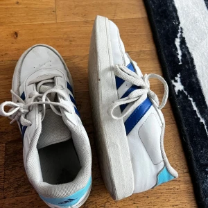 Vita Adidas sneakers med blå ränder - Klassiska vita Adidas sneakers med blå ränder på sidorna och ljusblå detalj vid hälen. Skorna har snörning, platt sula och rund tå. Tillverkade i syntetmaterial och skinnliknande ovandel. Perfekta för en sportig och avslappnad stil.