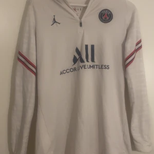 Vit långärmad PSG x Jordan fotbollströja - Snygg vit långärmad PSG x Jordan fotbollströja med halv dragkedja och huva. Tröjan har blå och röda ränder på ärmarna, PSG-logga på bröstet och Jordan-logga. Materialet är lätt och sportigt, perfekt för träning eller match. Tryck på ryggen och ärmen.