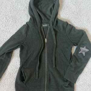 Mörkgrön hoodie från Zadig & Voltaire - Mörkgrå stickad hoodie från Zadig & Voltaire i 100 % kashmir med dragkedja framtill och huva. På båda armbågarna finns silverfärgade stjärnor. Köpte i affären i Paris för 2400 kr. Kan sälja för 700 vid snabb affär!!