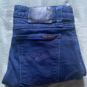 Mörkblå jeans från Nudie Jeans, W30 L30 - Snygga mörkblå jeans från Nudie Jeans med klassisk femficksdesign och diskret broderad bakficka. Jeansen har raka ben och är tillverkade i ett mjukt och slitstarkt bomullstyg. Perfekta för dig som gillar stilrena och tidlösa jeans.