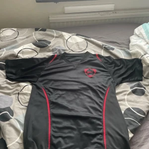 Svart Nike fotbollströja med röd detalj - Snygg svart fotbollströja från Nike med röda och vita detaljer längs sidorna och axlarna. Tröjan har V-ringad hals och korta ärmar, tillverkad i ett lätt och ventilerande material som passar perfekt för träning eller match. Nike-logga broderad på bröstet.