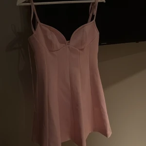 Ljusrosa kort klänning från Zara - Supersöt ljusrosa kort klänning från Zara med smala axelband och markerad byst. Klänningen har en lätt utsvängd form och dragkedja i sidan för enkel påklädning. Perfekt för dig som vill ha en feminin och trendig look.