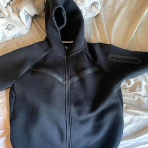 Svart Nike hoodiejacka XL - Svart hoodiejacka från Nike i storlek XL, med hel dragkedja framtill och huva. Jackan har en diskret Nike-logga på bröstet och en praktisk ficka med dragkedja på ärmen. Tillverkad i mjukt syntetmaterial, perfekt för chill eller träning.