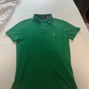 Grön pikétröja från Polo Ralph Lauren - Snygg grön pikétröja från Polo Ralph Lauren i custom slim fit. Klassisk krage, två knappar och korta ärmar. Broderad logga i rosa på bröstet. Perfekt för dig som gillar stilrena och sportiga plagg.