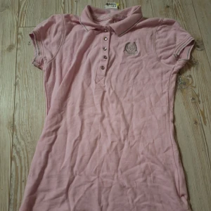 Rosa pikétopp med strass och tryck - Supersöt rosa pikétopp med glittriga strassknappar och silvrigt tryck både fram och bak. Topp med krage, korta ärmar och dekorativa detaljer som ger en sportig och söt vibe. Passar även som en kort klänning eftersom att den är relativt lång.