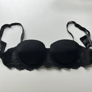 Svart bandeau-bh från Calvin Klein - Snygg svart bandeau-bh från Calvin Klein med avtagbara axelband och spetsdetaljer längs nederkanten. Tillverkad i mjuk nylonblandning med elastan för extra komfort och passform. Perfekt under toppar med bara axlar. Lätt push upp. 