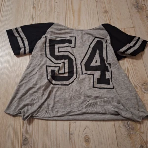 Grå jersey t-shirt med tryck - Snygg grå t-shirt från Bik Bok med svarta ärmar och stora siffran 54 tryckt på framsidan. Ärmarna har coola ränder och tröjan lutar mer mot en magtröja än en vanlig tröja. Perfekt för en sportig och chill stil.