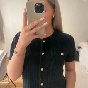 H&M Svart stickad topp med guldfärgade knappar - Säljer en svart topp med korta ärmar och rund hals. Toppen har snygga guldfärgade knappar framtill och två dekorativa fickor på bröstet. Perfekt för dig som gillar klassisk stil med en twist. Den är i Storlek L från H&M men passar S-L 