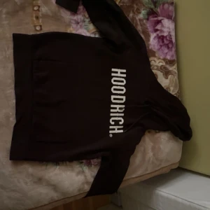 Svart HOODRICH hoodie med tryck - Svart hoodie från HOODRICH med stor vit logga tryckt vertikalt på framsidan. Klassisk känguruficka, huva med snörning och långa ärmar. Perfekt för en streetwear-look och chill dagar.