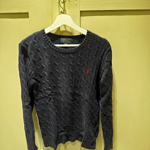  stickad tröja Polo Ralph Lauren - Kabelstickad mörkblå tröja från Polo Ralph Lauren med rund halsringning och klassisk röd broderad logga på bröstet. Tröjan har långa ärmar och ribbade muddar, perfekt för lager-på-lager-stil under kyliga dagar.