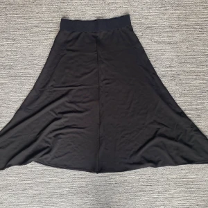 Svart långkjol från Tiger of Sweden - Svart långkjol från Tiger of Sweden, modell MABLE. Kjolen har en bred, ribbad midja och är lätt utsvängd med mjukt fall. Materialet är slätt och känns bekvämt mot huden, perfekt för dig som gillar stilrena och enkla plagg.