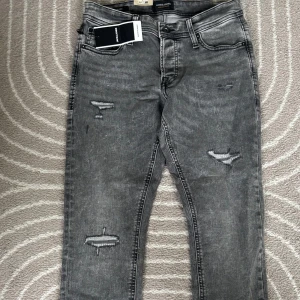 Nya Grå slim jeans från Jack & Jones - Snygga grå jeans från Jack & Jones i modellen Slim Glenn. Jeansen har slitna detaljer på benen och klassisk femficksdesign. De är slim fit och tillverkade i stretchig denim för en bekväm passform. Perfekta för en cool och avslappnad stil. Köpta tidigare för 900kr men ej använda så säljer för 50% även fast lappen är kvar.