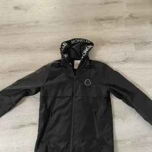 Svart vindjacka från Moncler - Svart vindjacka från Moncler med huva och vit Moncler-logga på bröstet. Huva med tydlig Moncler-tryck längs kanten. Jackan har dragkedja framtill och två fickor med dragkedja. Perfekt för blåsiga dagar och snygg streetstil.