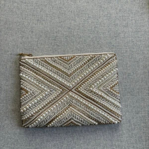 Pärlbroderad kuvertväska i satin - Superfin kuvertväska i ljus satin med pärl- och guldbroderier i geometriskt mönster på framsidan. Väskan har dragkedja upptill och insidan är fodrad i blankt tyg. Perfekt när du vill addera lite extra bling till din look.