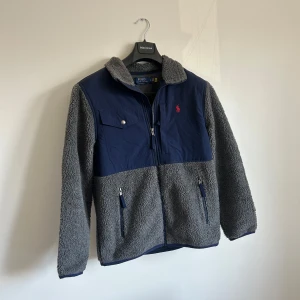 Grå och marinblå teddyjacka Polo Ralph Lauren - Säljer en grå teddyjacka från Polo Ralph Lauren med marinblå detaljer på bröst, axlar och armbågar. Jackan har dragkedja framtill, två fickor med dragkedja och en bröstficka med knapp. Perfekt för kyliga dagar och riktigt skön att ha på sig. Väldigt fint skick inte använd så mycket. Pris prutbart vid snabb affär 