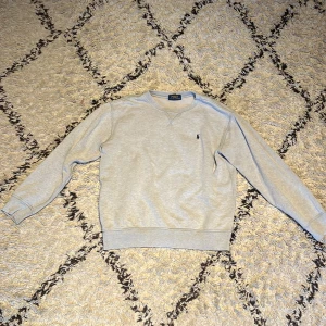 Grå sweatshirt från Polo Ralph Lauren - Klassisk grå sweatshirt från Polo Ralph Lauren med broderad marinblå logga på bröstet. Tröjan har rund halsringning, långa ärmar och ribbade muddar. Tröjan är i strlk L ( 14-16 )
