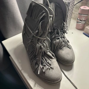 Grå mockaboots med fransar - Säljer ett par grå boots i mocka med massor av fransdetaljer längs skaftet och framtill. Skorna har rund tå, platt sula och snörning. Perfekta för dig som vill sticka ut med en cool och bohemisk stil.