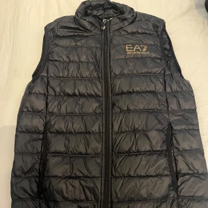 Svart dunväst från EA7 Emporio Armani - Snygg svart dunväst från EA7 Emporio Armani med quiltad design och dragkedja framtill. Västen har hög krage och diskret logga i guld på bröstet. Perfekt nu till hösten. Skick 10/10 knappt använd. Storlek M. Köpt för 1299kr på Zalando. Säljer för 400kr. Pris kan diskuteras vid snabb affär. Vid fler frågor eller intresse skriv!