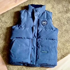 Mörkblå dunväst från Canada Goose - Snygg mörkblå dunväst från Canada Goose med hög krage och två stora fickor framtill. Västen har tryckknappar och ett broderat logomärke på bröstet. Perfekt för kyliga dagar och har en klassisk, quiltad look.