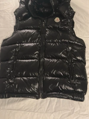 Svart dunväst från Moncler - Säljer en svart, glansig dunväst från Moncler med hög krage och två dragkedjeförsedda fickor framtill. Västen har klassisk Moncler-logga på bröstet och stängs med dragkedja. Perfekt för lager-på-lager under höst och vinter.