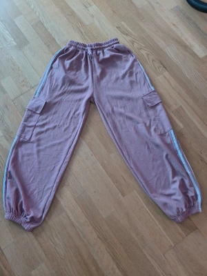 Ljuslila cargopants med vita ränder - Säljer ett par ljuslila cargopants med elastisk midja och muddar vid bensluten. Byxorna har två stora sidofickor med lock och vita ränder längs benen.  perfekta för chill dagar.