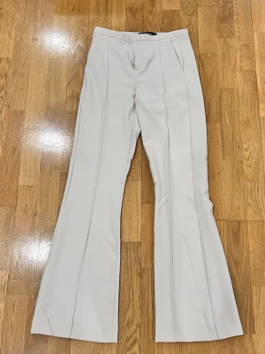 Beiga bootcut kostymbyxor ginatricot  - Snygga beiga kostymbyxor med bootcut passform och pressveck längs benen. Byxorna har hög midja och är tillverkade i ett mjukt, vävt material som ger en clean och stilren look. Perfekta för dig som vill ha en trendig siluett.