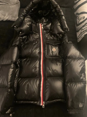 Svart Moncler jacka - Säljer en svart jacka från Moncler med glansig finish, huva och Moncler-logga på bröstet. Jackan har dragkedja med rödvit detalj och en praktisk bröstficka. Perfekt för kalla dagar. Villig att diskutera priset vid snabb affär.