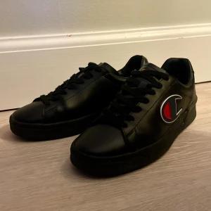Svarta sneakers från Champion - Säljer ett par svarta sneakers från Champion med stort logomärke i rött, vitt och blått på sidan. Skorna har snörning, rund tå och platt sula. Tillverkade i syntetmaterial med en clean och stilren look som passar till det mesta.