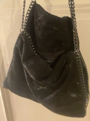 Svart axelväska med kedjedetaljer - Säljer en svart axelväska med glansig finish och grova silverfärgade kedjor längs kanterna och som axelband. Väskan har en mjuk, slouchy form och känns riktigt trendig. Perfekt för dig som vill ha en statementväska med coola detaljer.